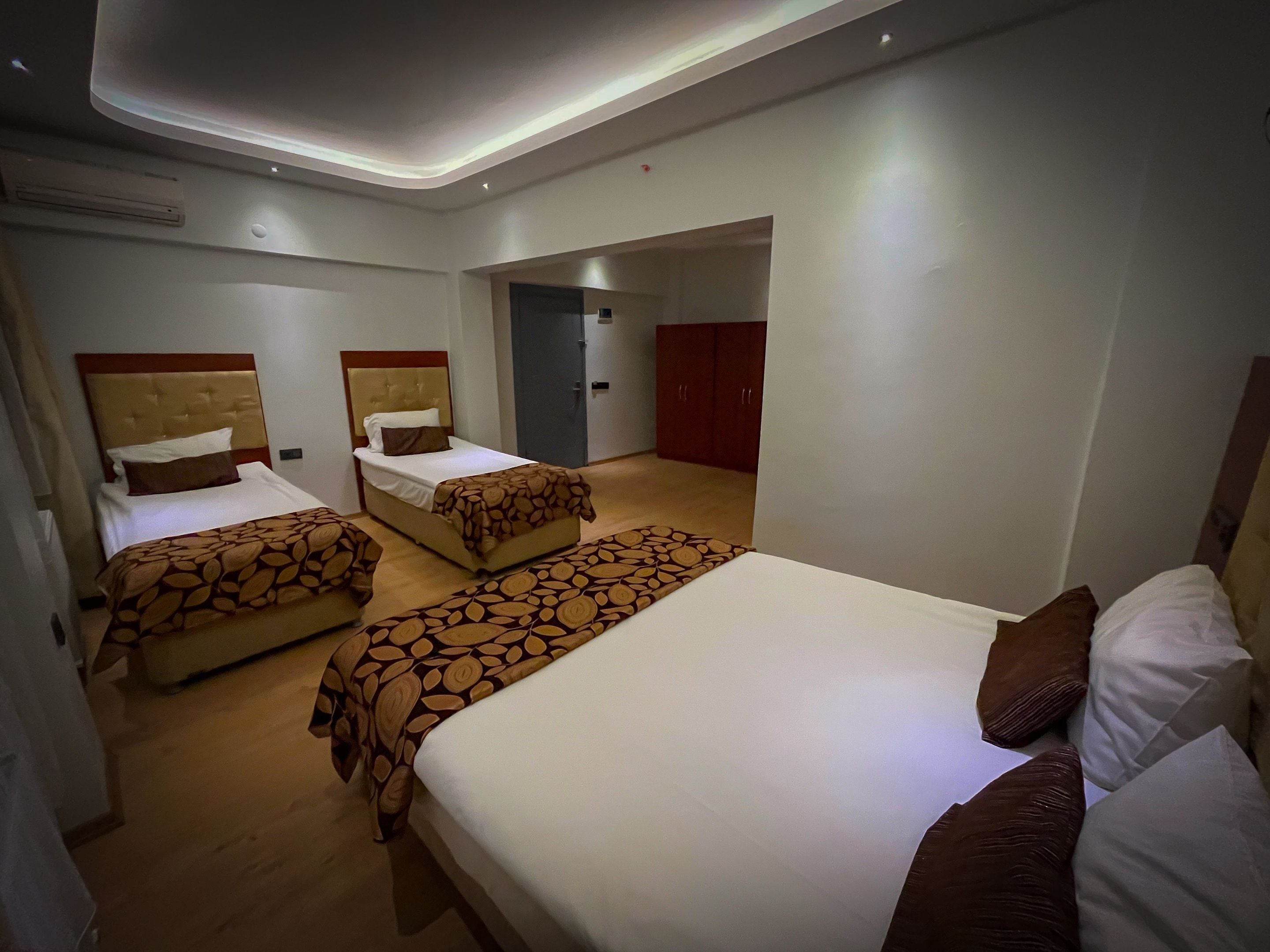 Une chambre d’hôtel avec trois lits, chacun recouvert d’une couvre-lit à motifs, doté d’une tête de lit en bois et agrémenté de linge de lit blanc. On y trouve également une armoire et une porte donnant sur une autre pièce.