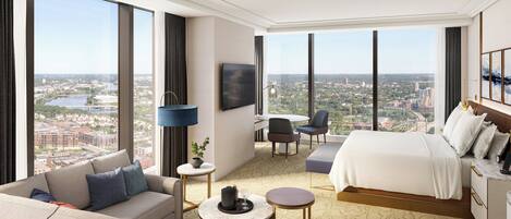 A modern hotel room with a large bed, a sofa, a dining table, and a TV. The room has large windows offering a city view.