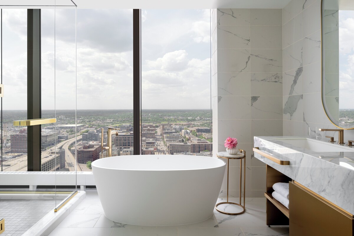Une salle de bain moderne avec une baignoire sur pieds, un grand miroir et une vue sur le paysage urbain, visible à travers une grande fenêtre.