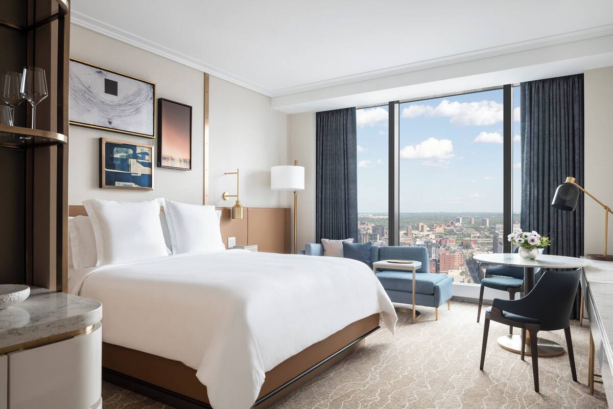 Een hotelkamer met een groot bed, een bureau met twee stoelen, uitzicht op de stad door grote ramen, en een stadsgezicht buiten.