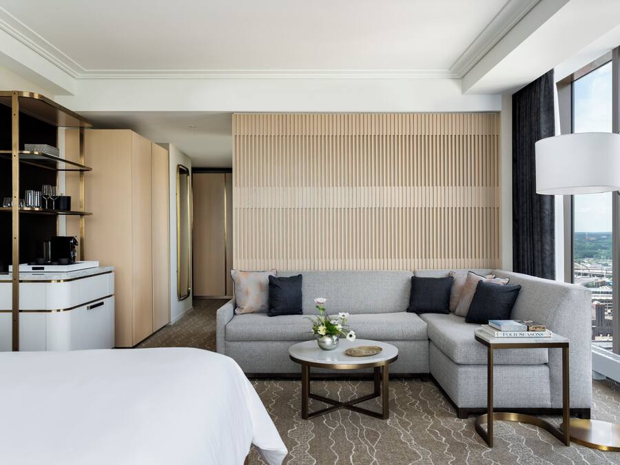 A modern hotel room with a bed, sofa, and a small table with a vase. There is a wardrobe, a lamp, and a view of the city.