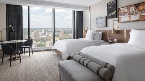 A hotel room with two beds, a desk, chairs, and a large window with a city view.