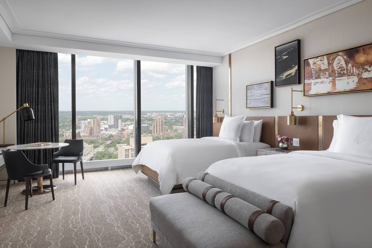 Een hotelkamer met twee bedden, een bureau, stoelen en een groot raam met uitzicht op de stad.