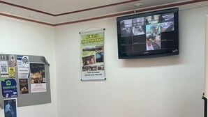 A waiting area with two black leather chairs, a small table with magazines, a poster on the wall, and a mounted TV displaying multiple camera feeds.
