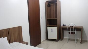 Quarto com cama de madeira, guarda-roupa, escrivaninha com cadeira e um pequeno frigorífico.
