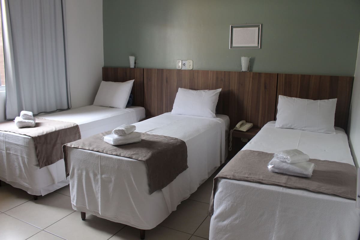 Une chambre d’hôtel avec trois lits simples, chacun recouvert d’une courtepointe blanche et d’un plaid marron. On y trouve une tête de lit en bois, un tableau encadré au mur et une table de chevet avec un téléphone.