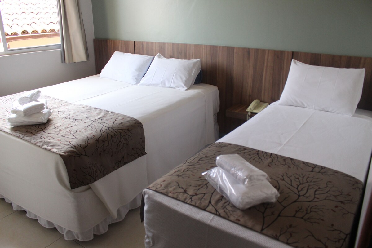 Two beds with white linens, a wooden headboard, a bedside table with a phone, and a window with curtains.