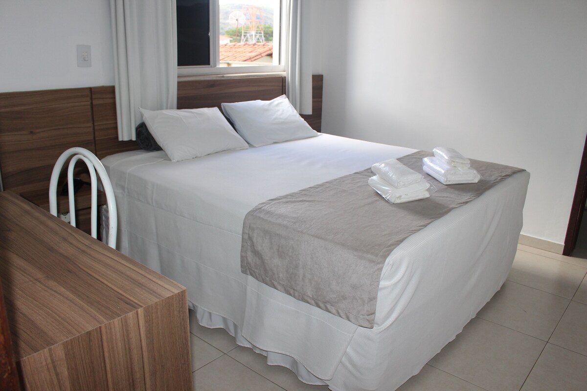A hotel room with a bed, a wooden bedside table, a window with curtains, and a view of the outside.