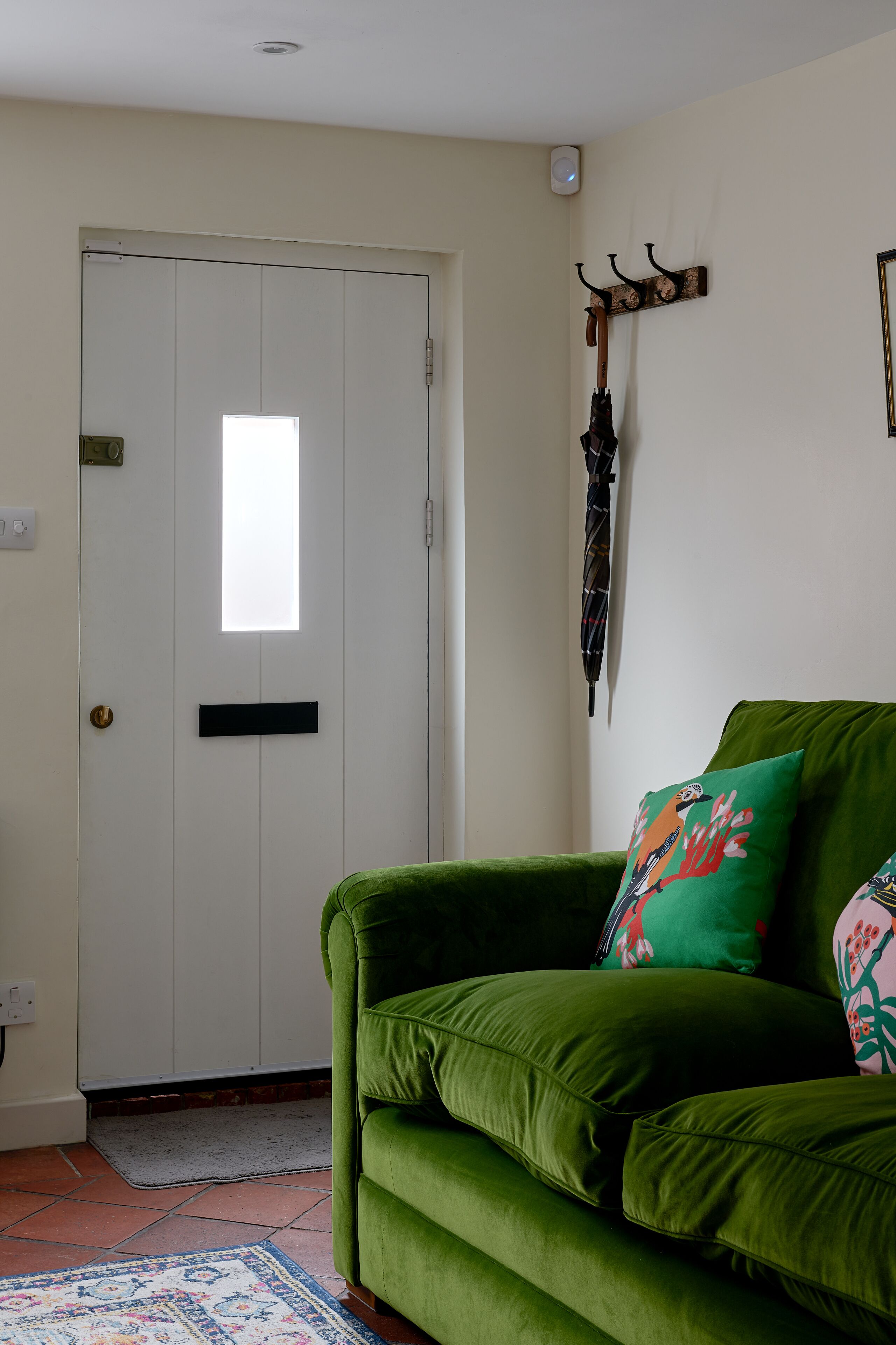 A green sofa with colorful cushions, a white door with a window, and a wall-mounted coat rack with an umbrella.