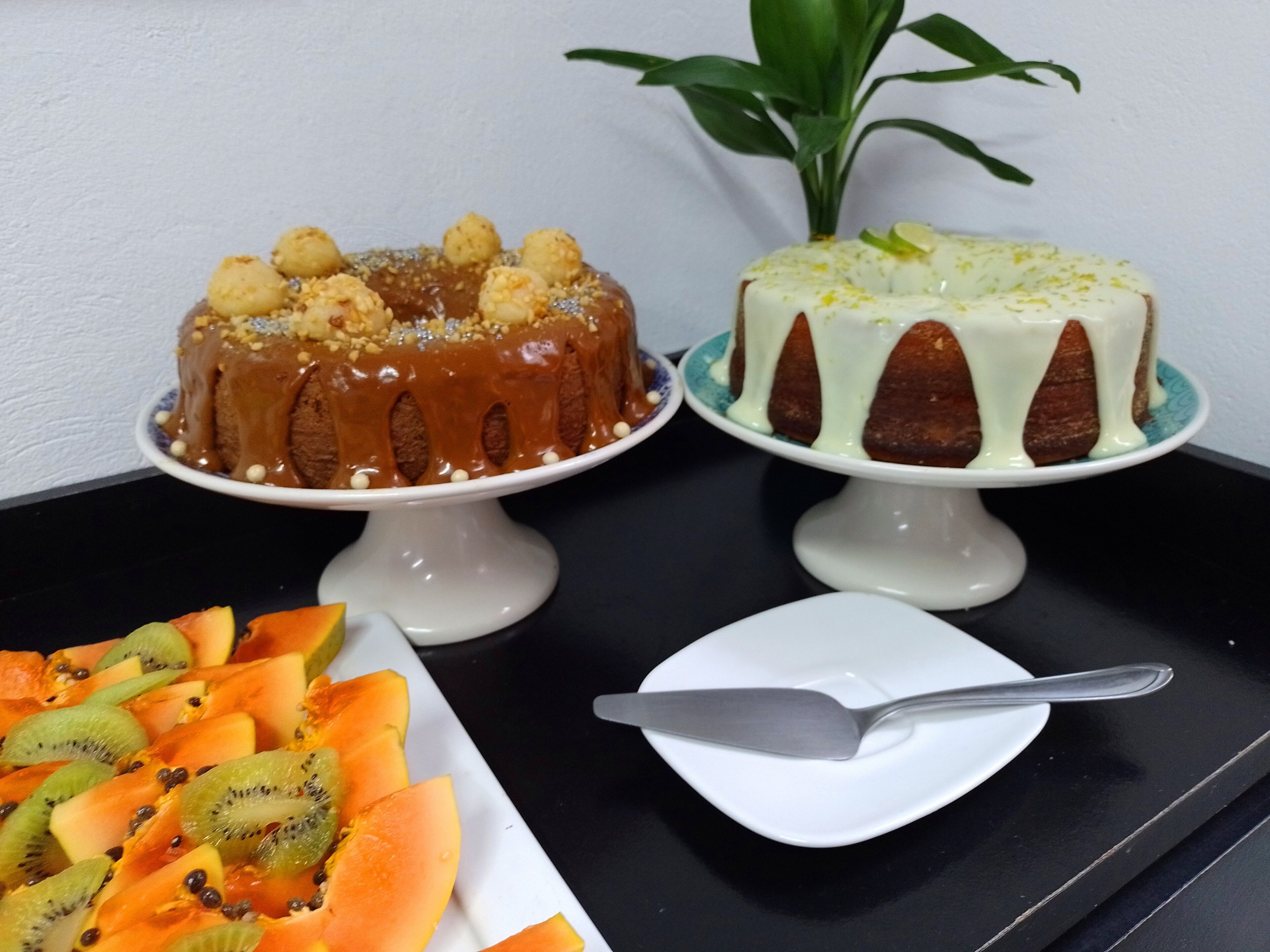 Two cakes on white stands with a plate of sliced fruit and a knife on a black table.