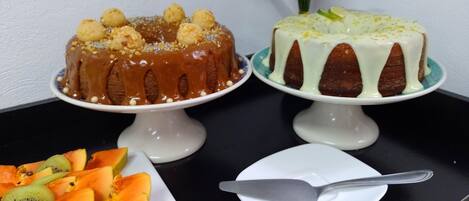 Two cakes on white stands with a plate of sliced fruit and a knife on a black table.