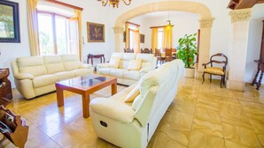 A spacious living room with a sofa, armchair, and coffee table, a dining area with chairs, and a large window with curtains.