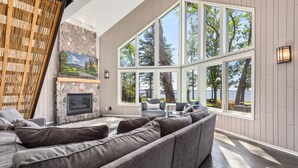 Un salon moderne doté d’une cheminée en pierre, de grandes fenêtres et offrant une vue sur les arbres et l’eau.