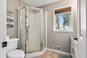 Un baño con cabina de ducha de vidrio, inodoro, ventana con vistas a árboles y un perchero de toallas en la pared.