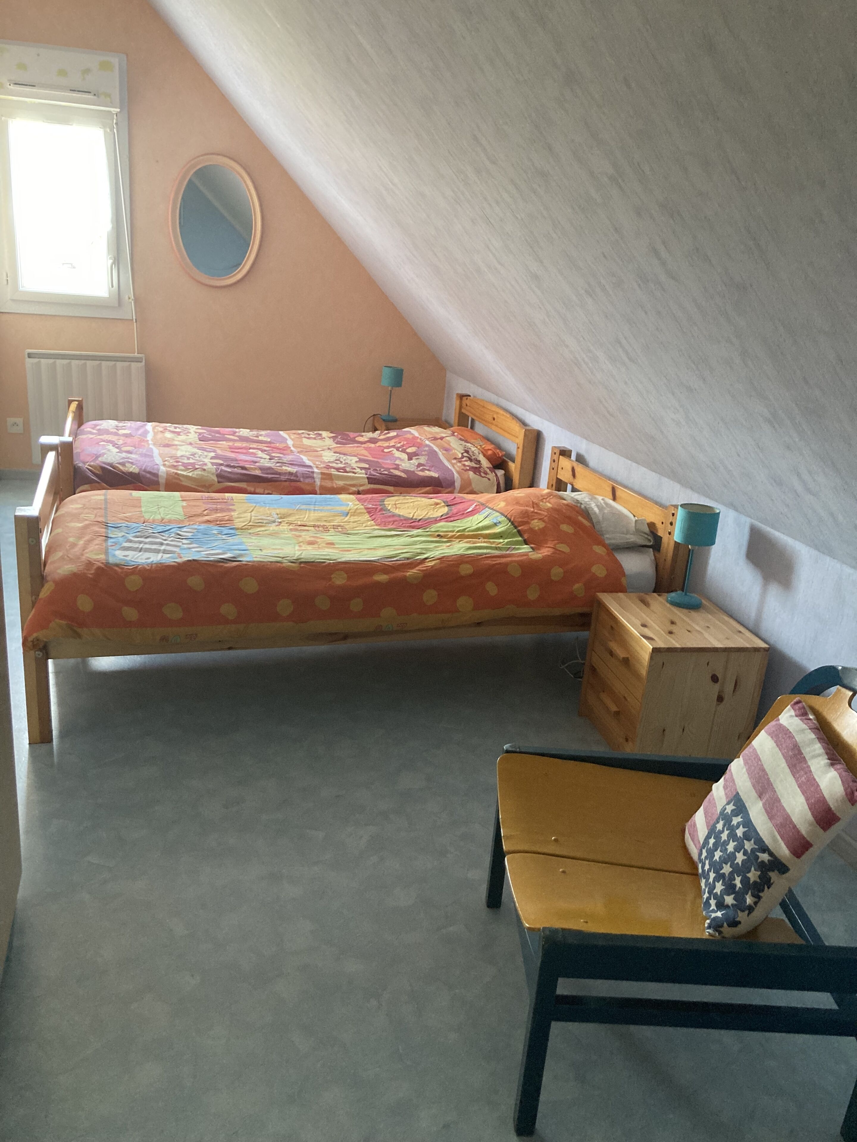 A small, cozy attic bedroom with two wooden beds, a small wooden chest, and a chair with an American flag pillow.