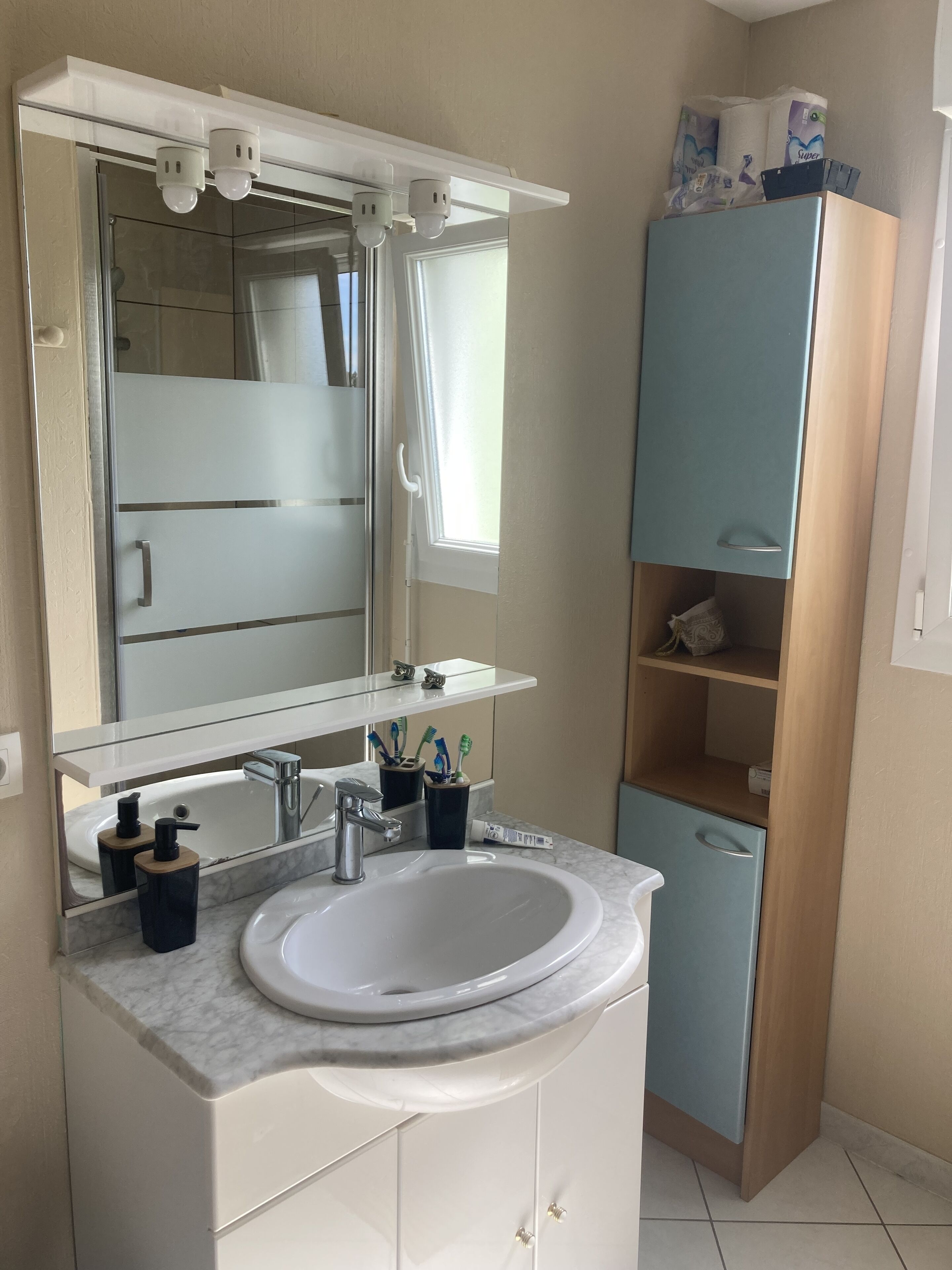 Ein Badezimmer mit einem weißen Waschbecken, einem Spiegel, einem blauen Schrank und einem Fenster.