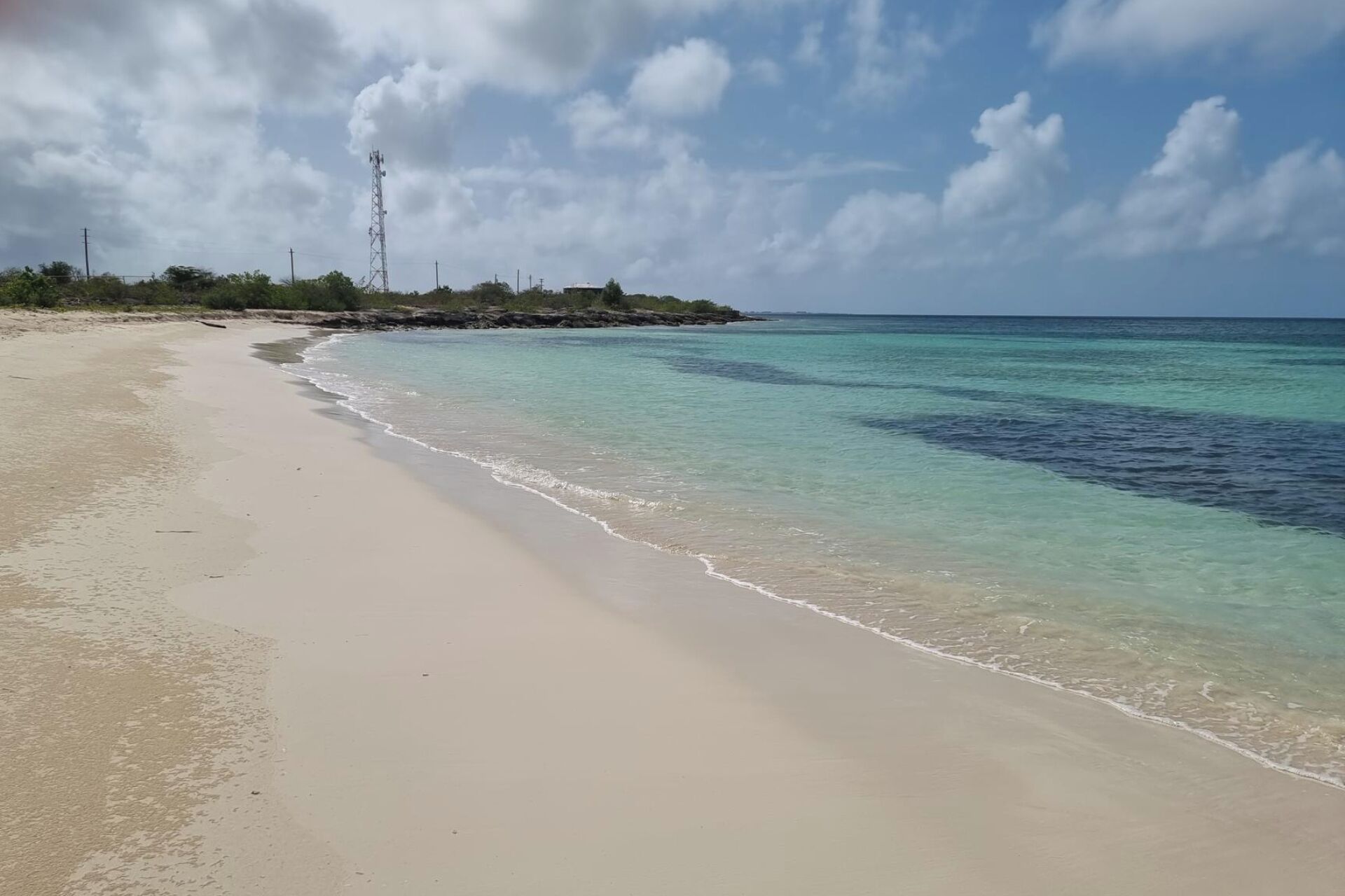 Una playa con agua turquesa cristalina, orilla arenosa y una torre de tendido eléctrico a lo lejos.