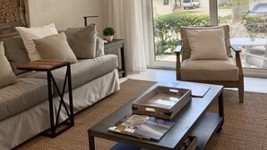 Salón con sofá, sillones, una mesita de centro con revistas y vista a una zona residencial con casas y un vehículo.
