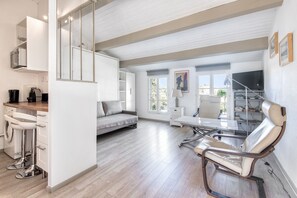 A modern living room with a sofa, a dining table, and a TV. There is a kitchen area with a sink and a coffee machine.