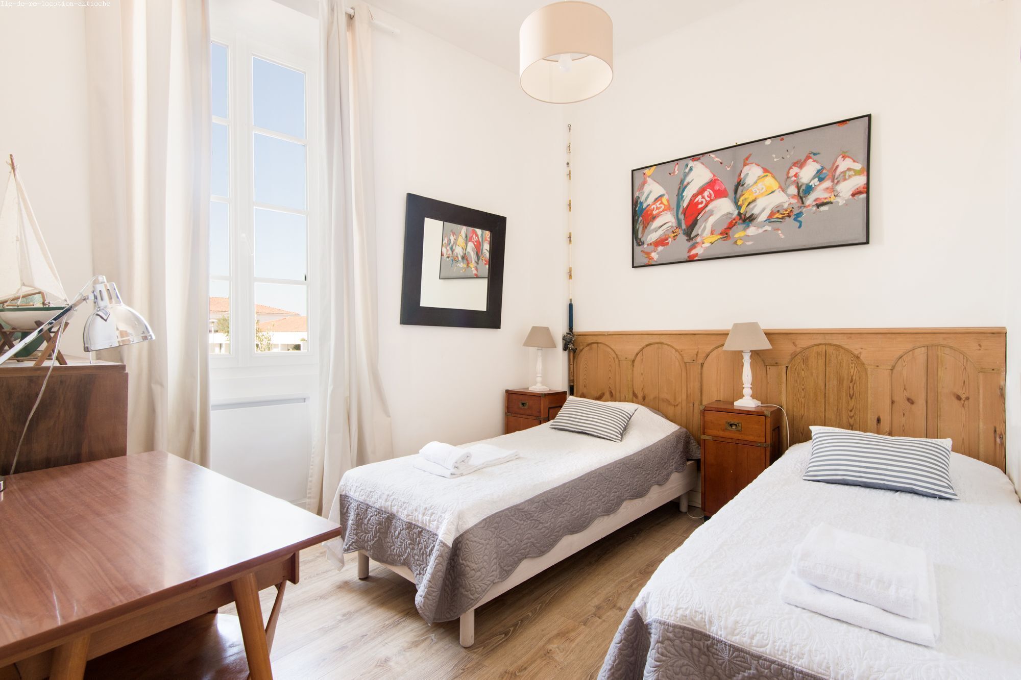 Habitación de hotel con dos camas individuales, cabecero de madera, escritorio, silla, lámpara, un cuadro enmarcado y una ventana con cortinas.
