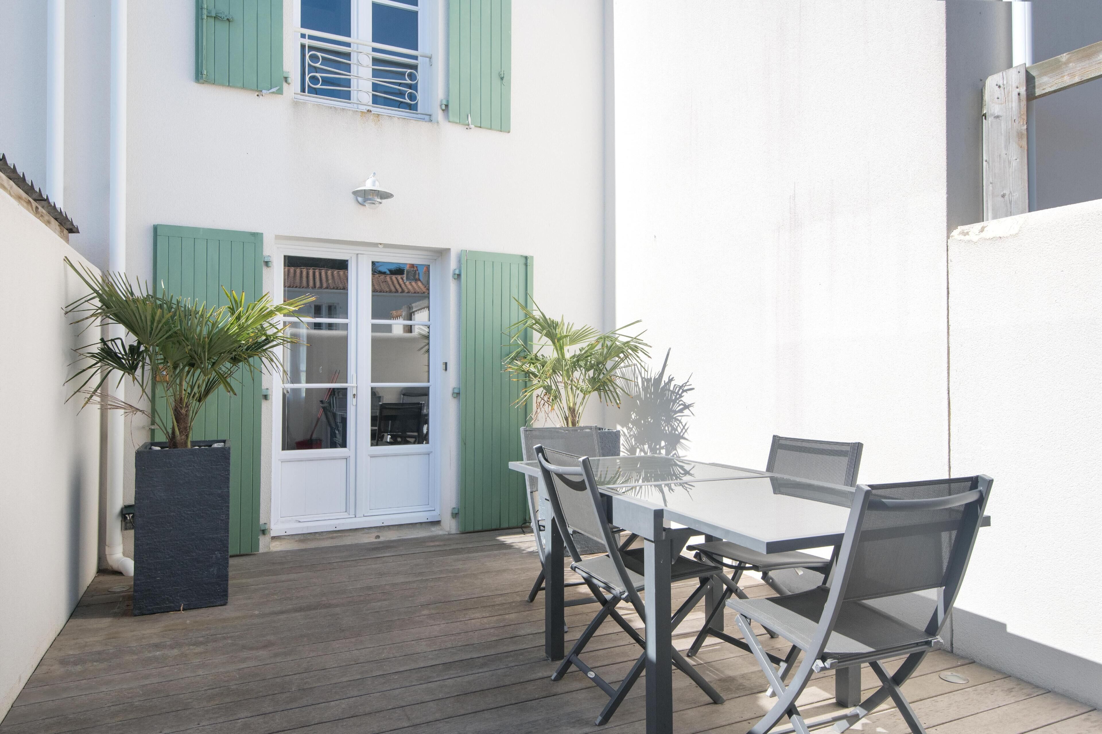 Une terrasse avec une table et des chaises, un pot de fleurs et une porte avec des volets verts.