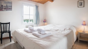 Ein ordentlich bezogenes Bett mit aufgerollten Handtüchern, ein Holzstuhl, ein Fenster mit Vorhängen, eine Lampe und ein gerahmtes Bild an der Wand.