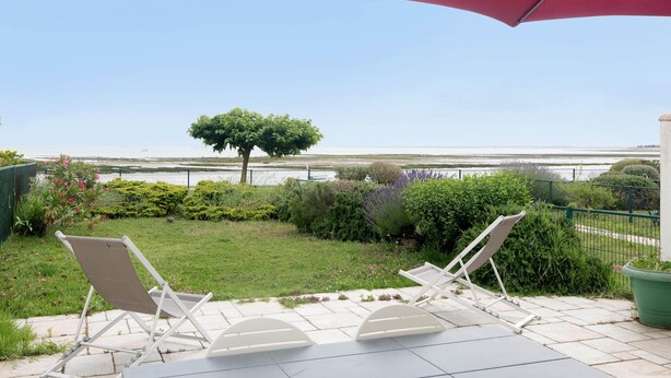Eine Terrasse mit zwei weißen Liegestühlen und einem roten Sonnenschirm mit Blick auf einen angelegten Garten und ein Gewässer.
