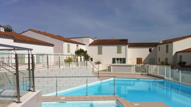 A swimming pool with a tiled deck, white buildings with green shutters, and a clear sky.