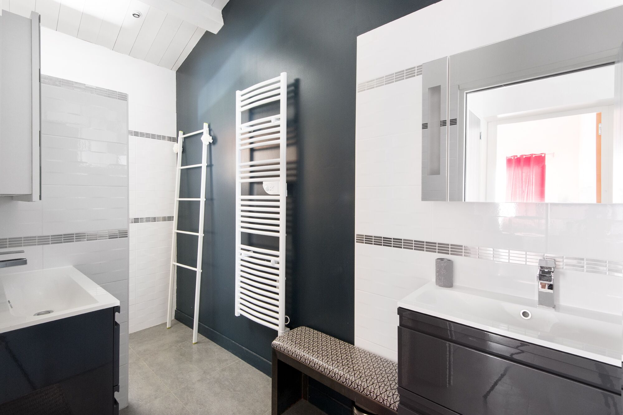 A modern bathroom with white and dark grey tiles, a white towel rack, a bench, and a large mirror.