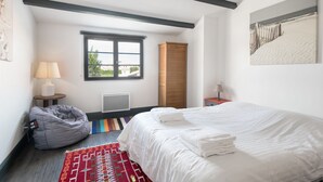 Un dormitorio con cama, ventana, radiador, armario, lámpara, sillón de mimbre y alfombra.