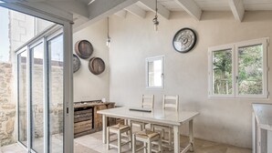 A dining area with a white table and chairs, wooden barrels on the wall, a clock, and a window with a view of greenery. - House SALICORNE    . (La Flotte)