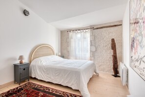 A bedroom with a bed, a nightstand, a lamp, a window with sheer curtains, a stone wall, and a wooden sculpture. - House SALICORNE    . (La Flotte)