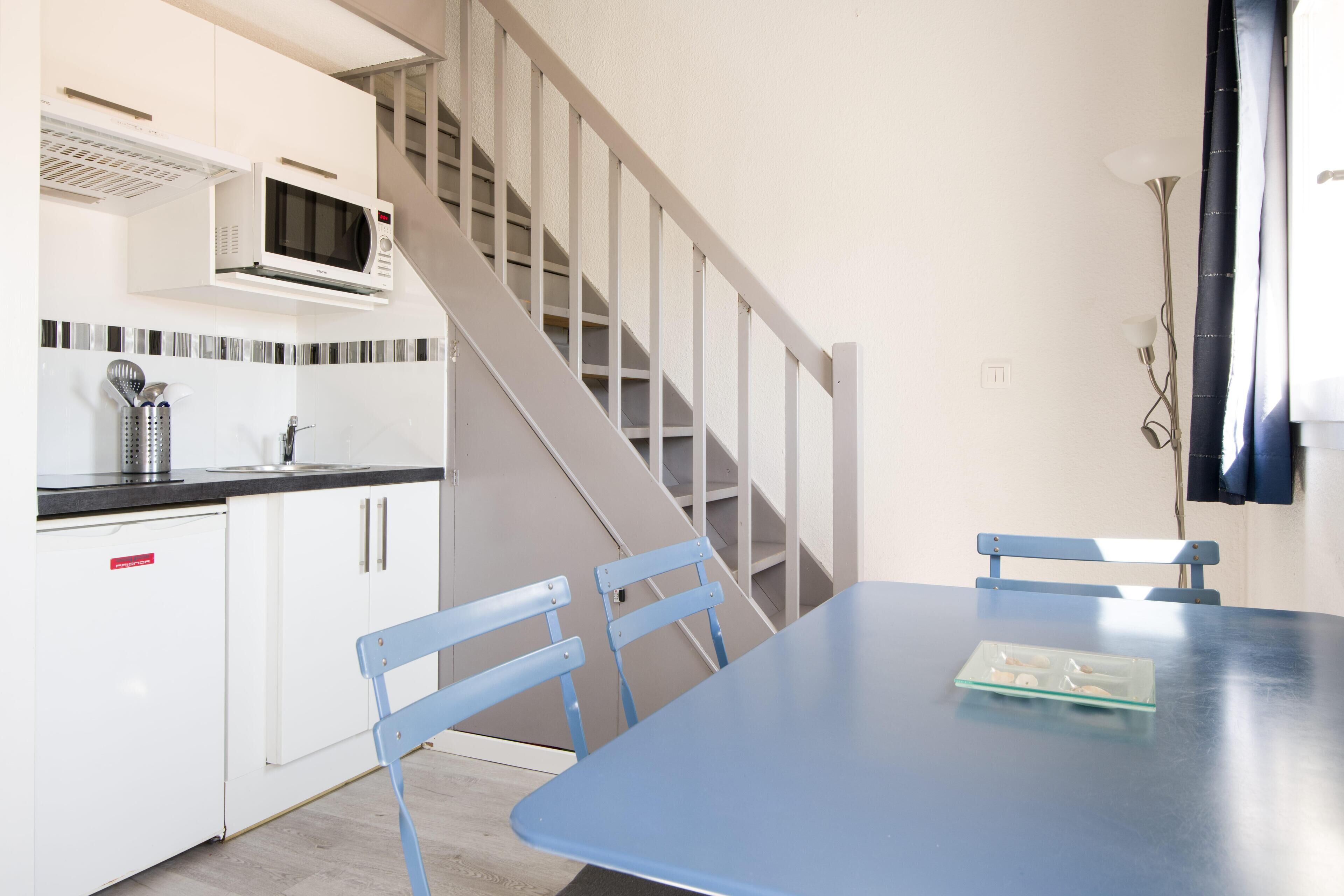 Una cocina con armarios blancos, un microondas y una escalera que lleva a otro nivel. Hay una mesa azul con sillas y una pequeña bandeja encima.