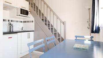Una cocina con armarios blancos, un microondas y una escalera que lleva a otro nivel. Hay una mesa azul con sillas y una pequeña bandeja encima.