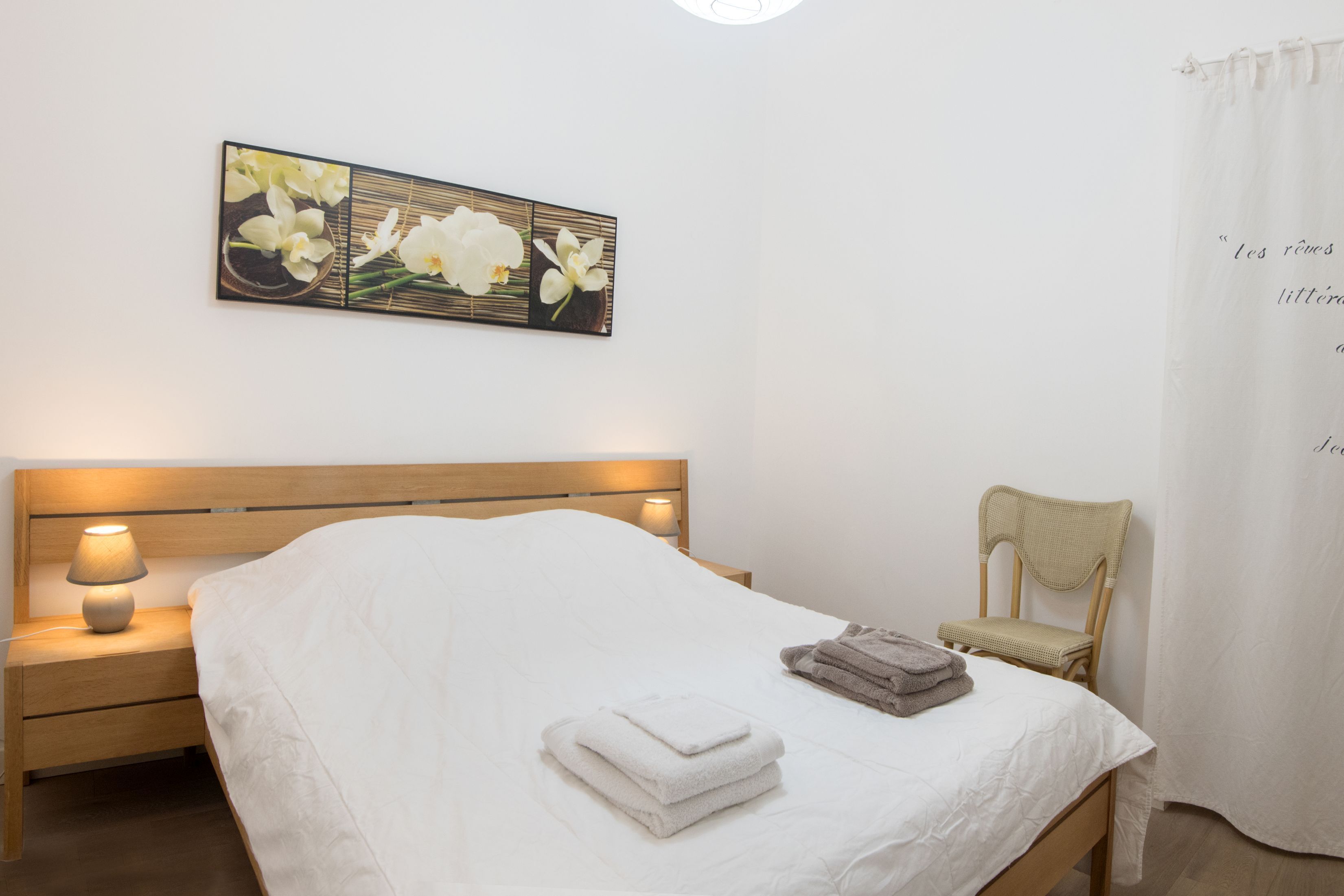 Una cama bien hecha con sábanas blancas, una lámpara de noche, una silla y una obra de arte en la pared con flores blancas.
