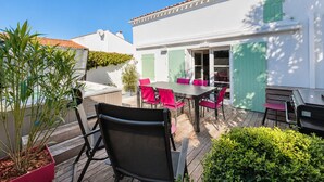 Une terrasse avec une table et des chaises, un pot de fleurs et un bâtiment blanc aux volets verts.