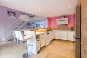 A modern kitchen with white cabinets, a built-in dishwasher, a stainless steel refrigerator, and a dining area with white chairs. - House AMANDE with private spa (La Flotte)