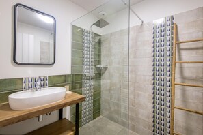 Une salle de bain avec un lavabo blanc, un miroir, une douche avec une porte en verre et un escabeau en bambou.