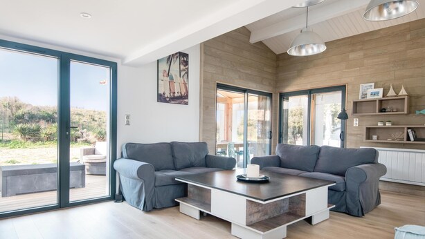 A modern living room with a grey sectional sofa, a glass coffee table, and a view of a garden through large sliding doors. - DUNE               . (La Couarde-sur-Mer)