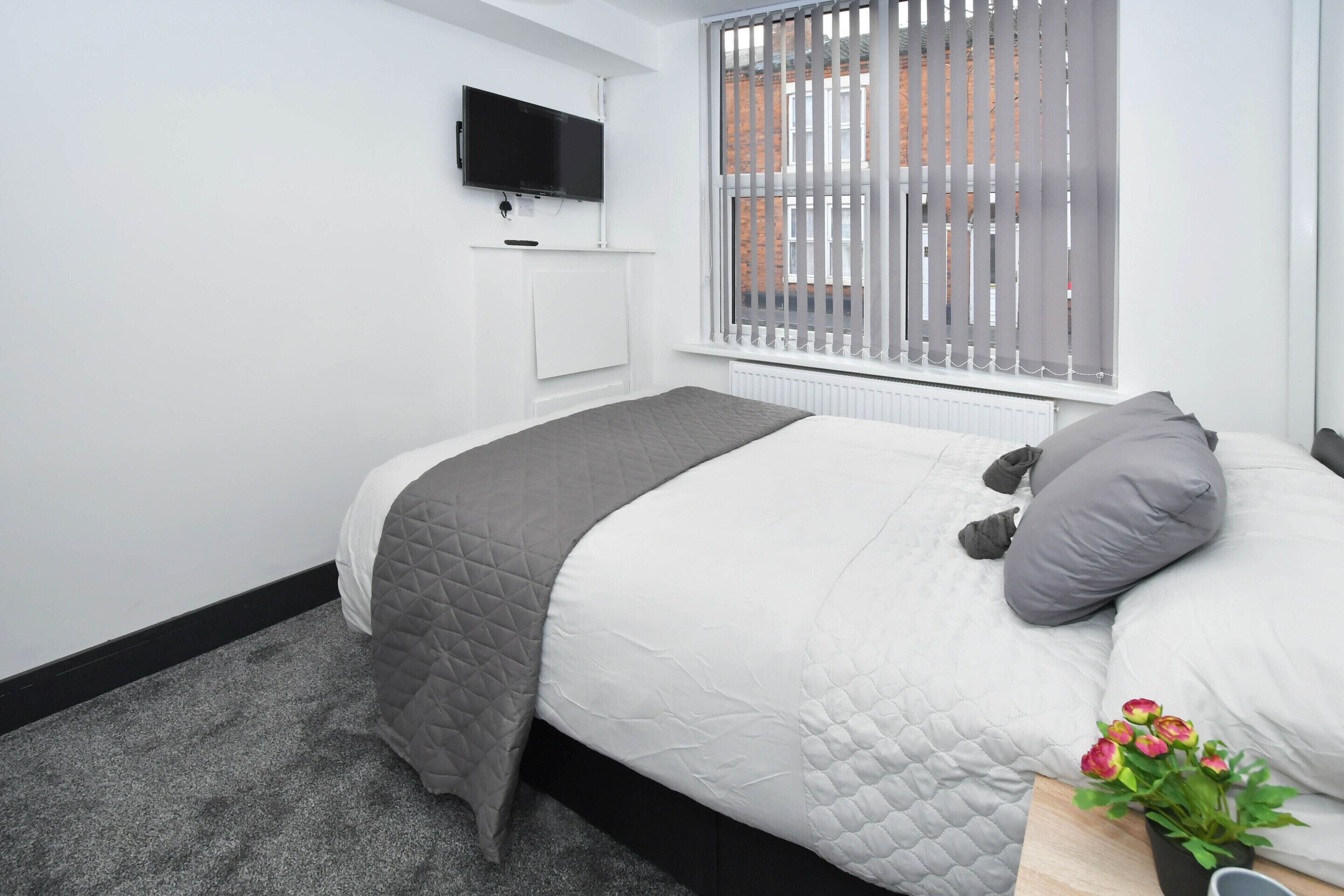 A bedroom with a bed, a TV mounted on the wall, a window with blinds, and a small table with a plant.