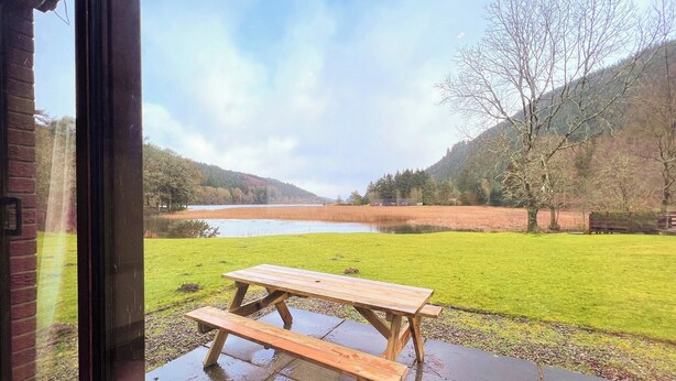 Ein hölzernes Picknicktisch mit Bänken auf einer feuchten Terrasse mit Blick auf einen See und Hügel.