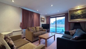 Ein Wohnzimmer mit Sofa, Sessel und Couchtisch. Ein Fernseher steht auf einem Schrank, ein Gemälde hängt an der Wand, und ein Fenster ist mit Vorhängen versehen.