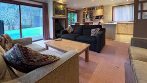 Ein Wohnzimmer mit Sofa, Sessel und Couchtisch. Ein Kamin, ein Bücherregal und ein Blick ins Freie sind ebenfalls vorhanden.