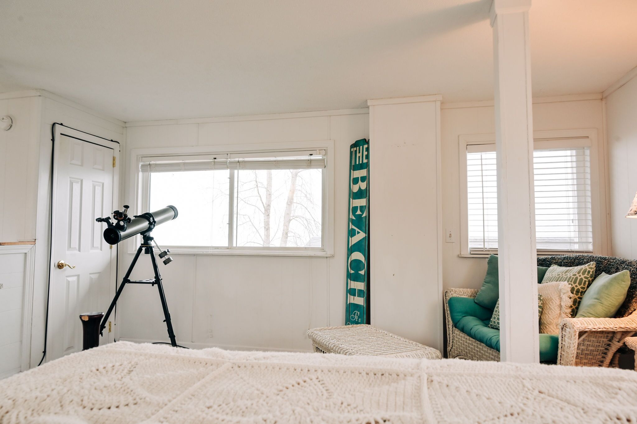 A bedroom with a telescope, a wicker chair, and a large window.