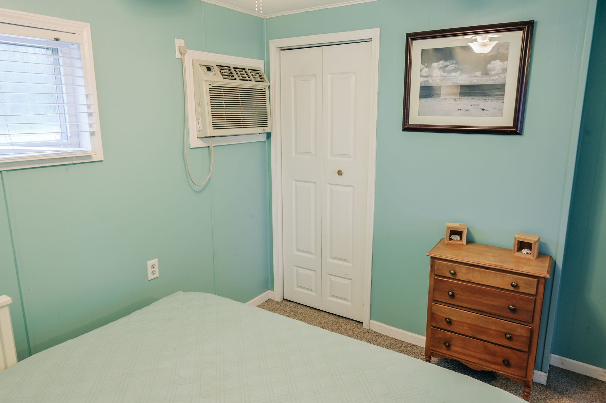 A bedroom with a single bed, a white door, a dresser, a framed picture on the wall, and an air conditioning unit.