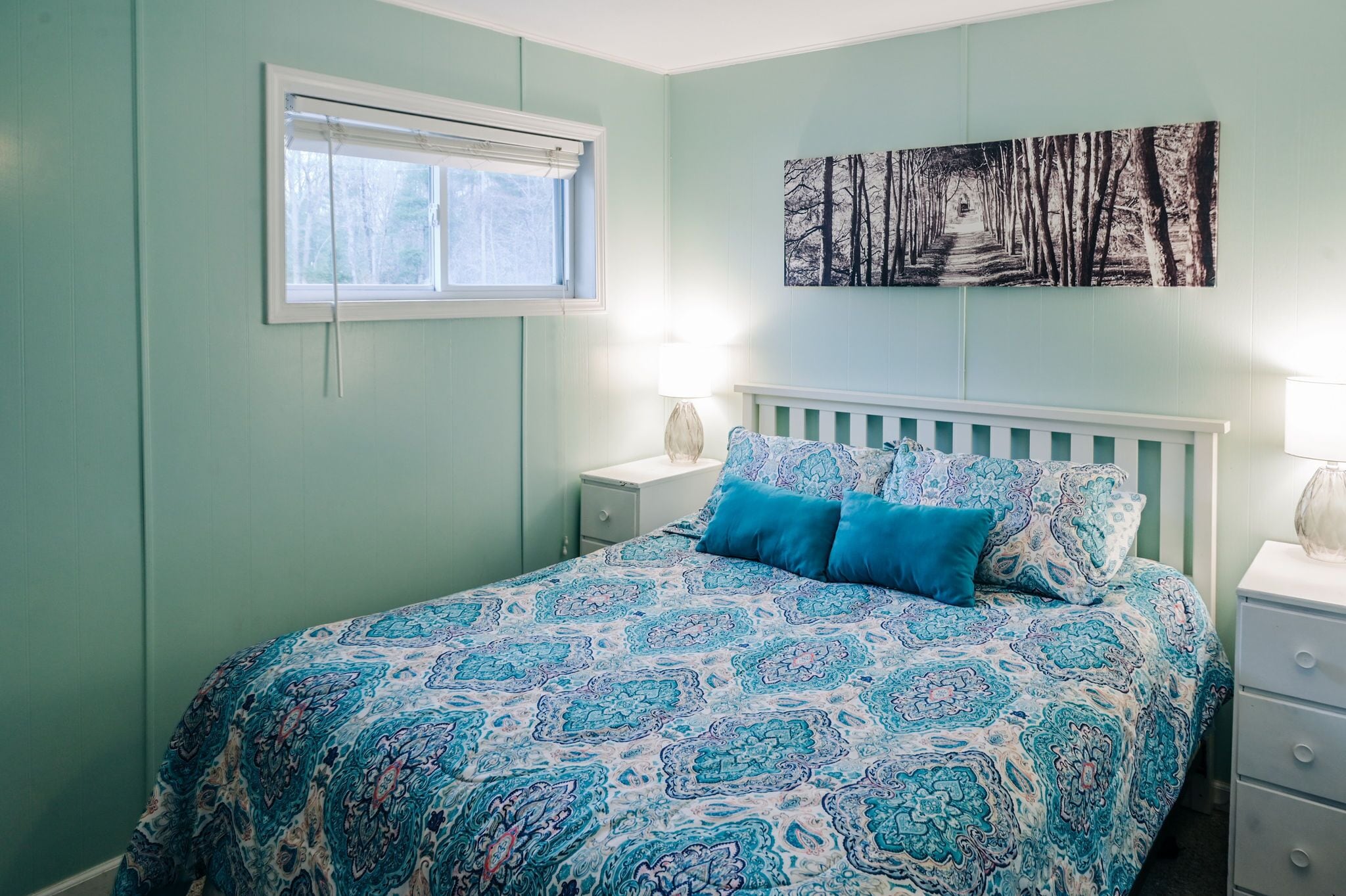 Een slaapkamer met een bed, nachtkastje, raam en een aan de muur hangend schilderij.