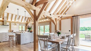 Una cocina amplia con vigas de madera, un comedor con sillas blancas y vistas a zonas verdes.