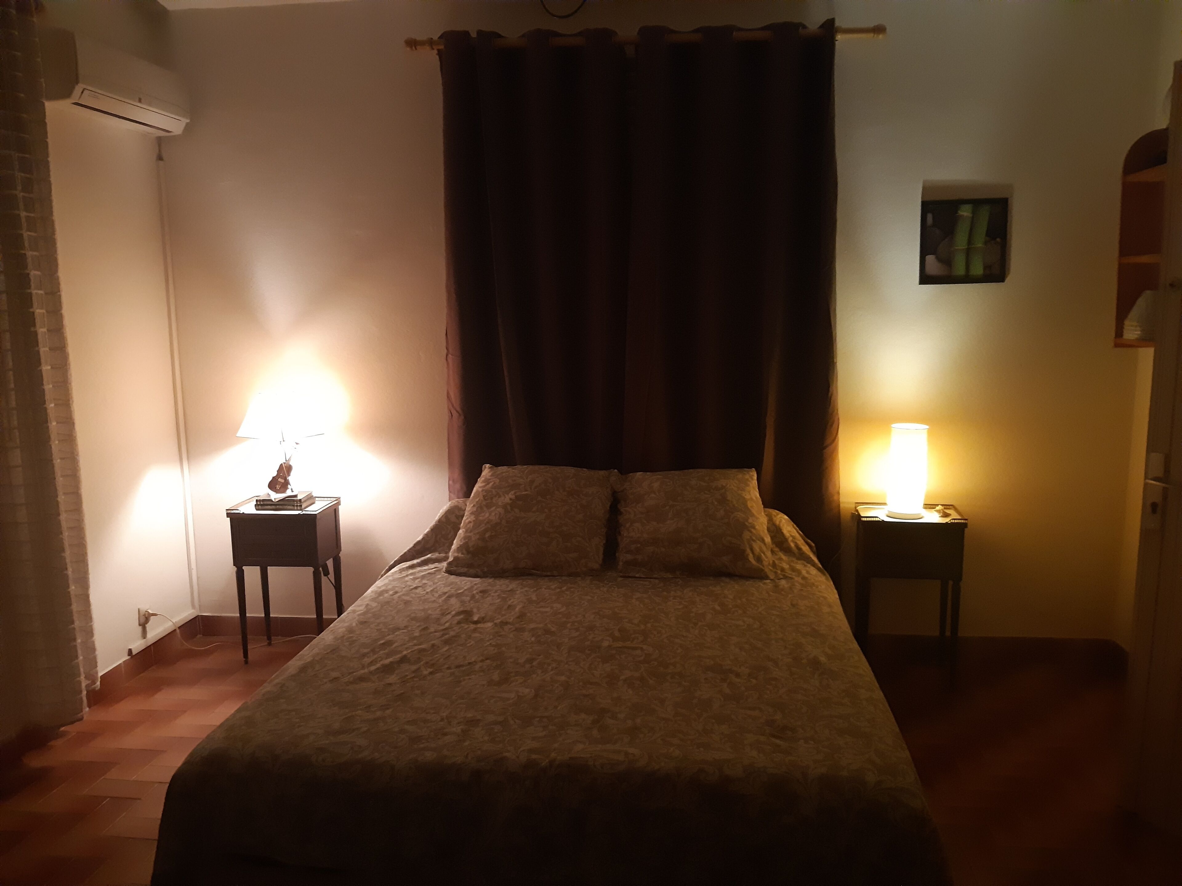Un dormitorio con una cama, dos lámparas de noche, un aire acondicionado instalado en la pared y un pequeño marco de fotos.