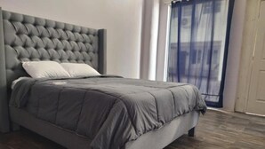 A modern bedroom with a tufted headboard, a grey bedspread, and a large window with a view of buildings.