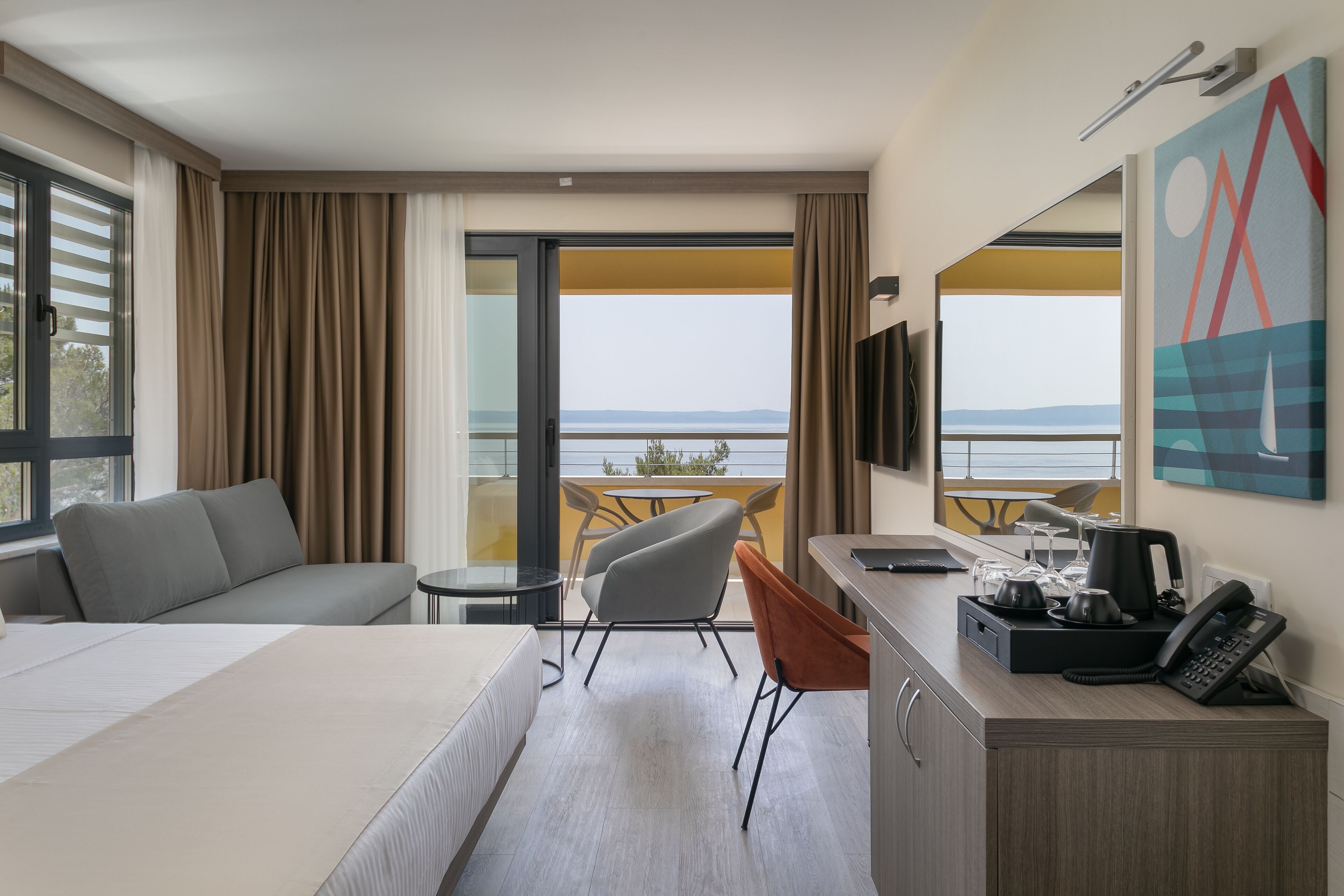 Una habitación de hotel con cama, escritorio con teléfono, una silla, un sofá y un balcón con mesa y sillas.